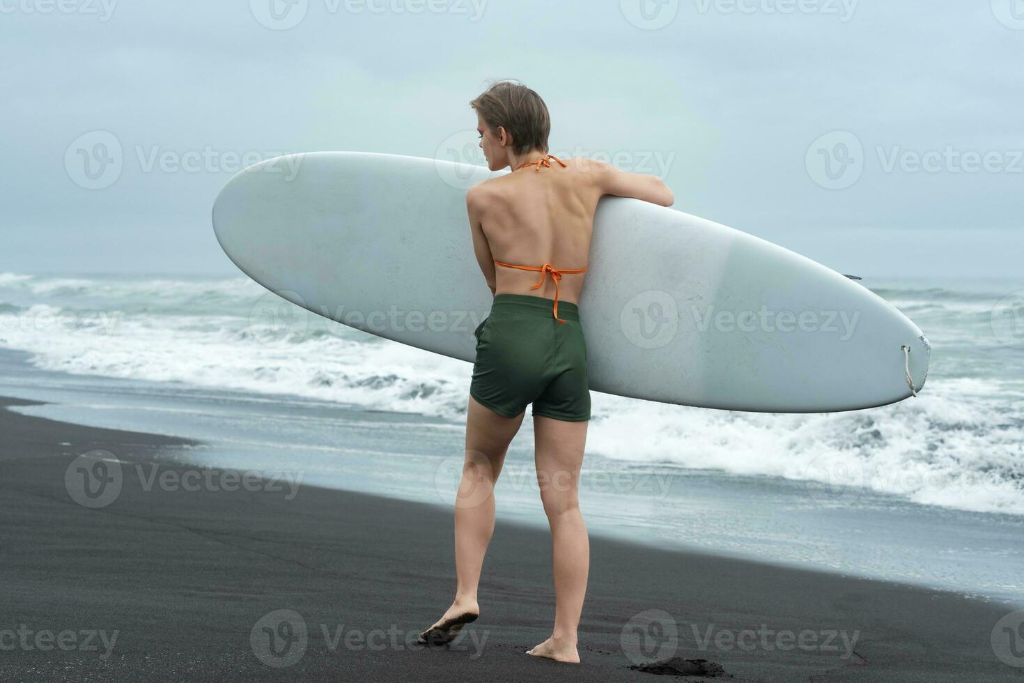 Rear view of female surfer walking on sandy beach carrying surfboard on background of ocean waves photo