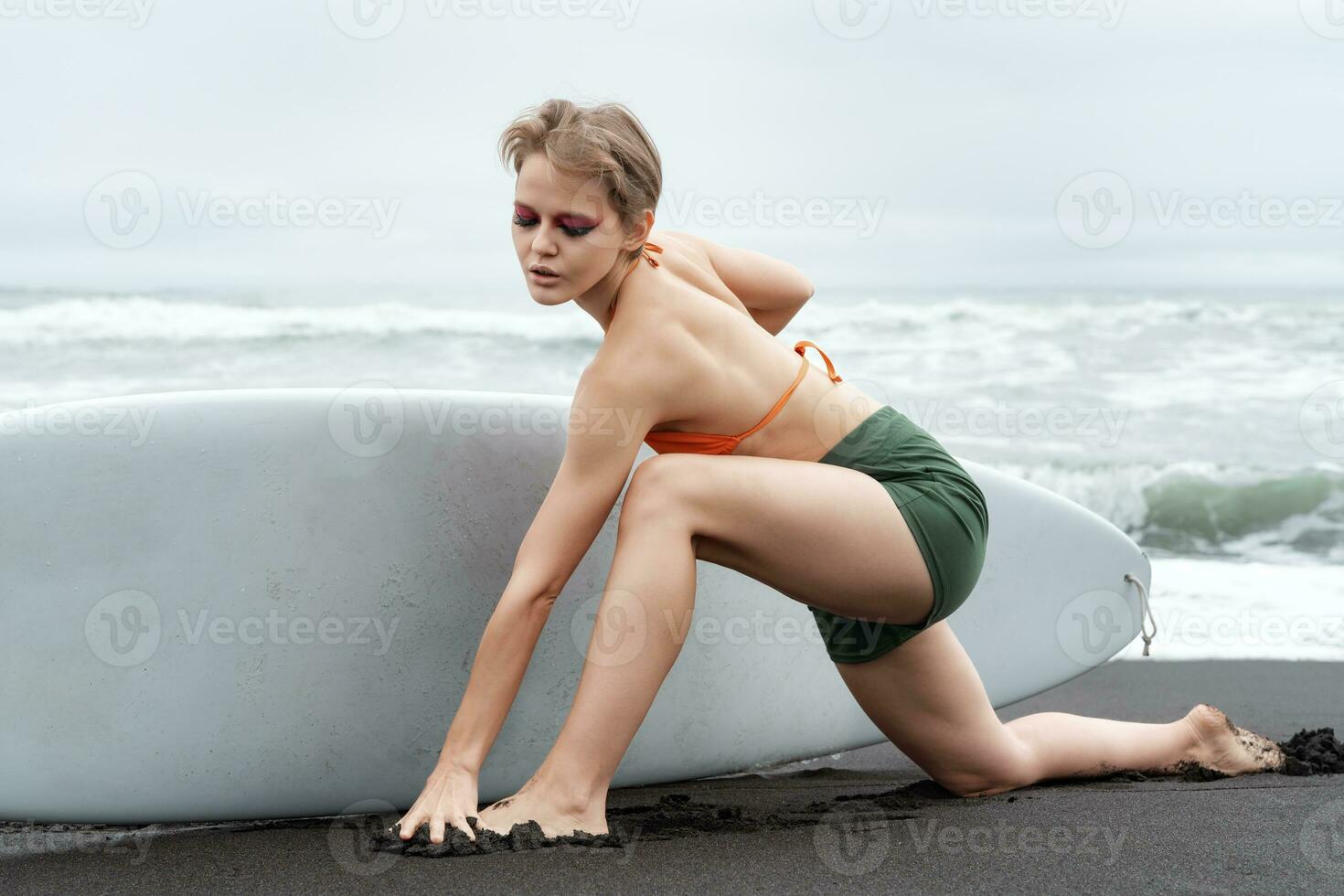 Female surfer stands on one knee, stretching leg forward on beach, and surfboard with one hand photo