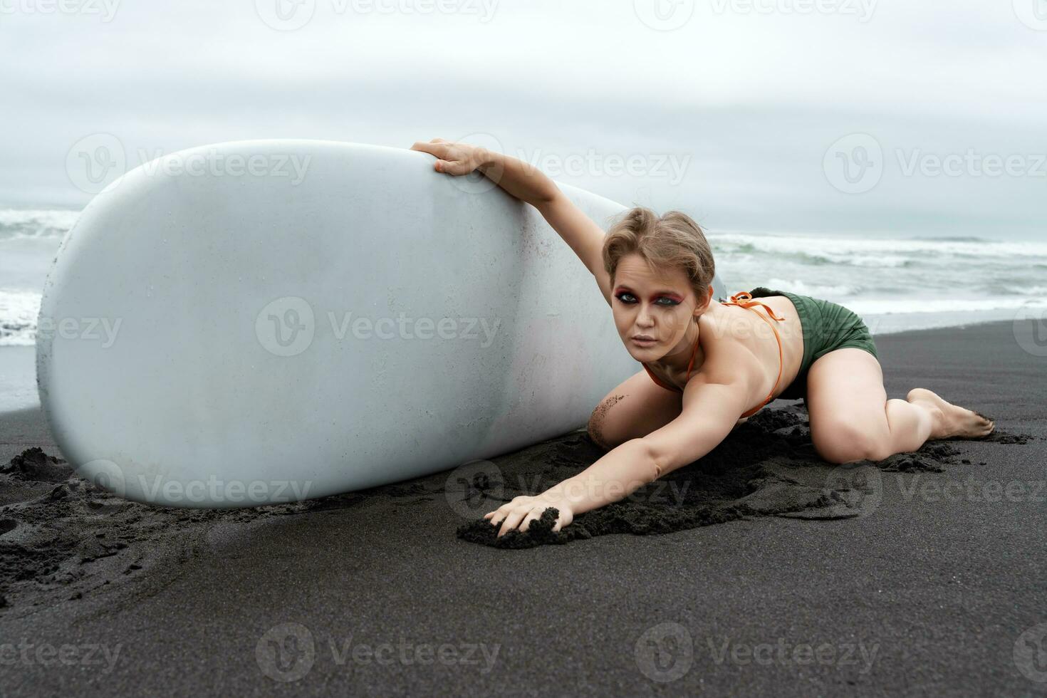 Amazing female surfer looks so cool kneeling on sand at beach, holding surfboard, looking at camera photo