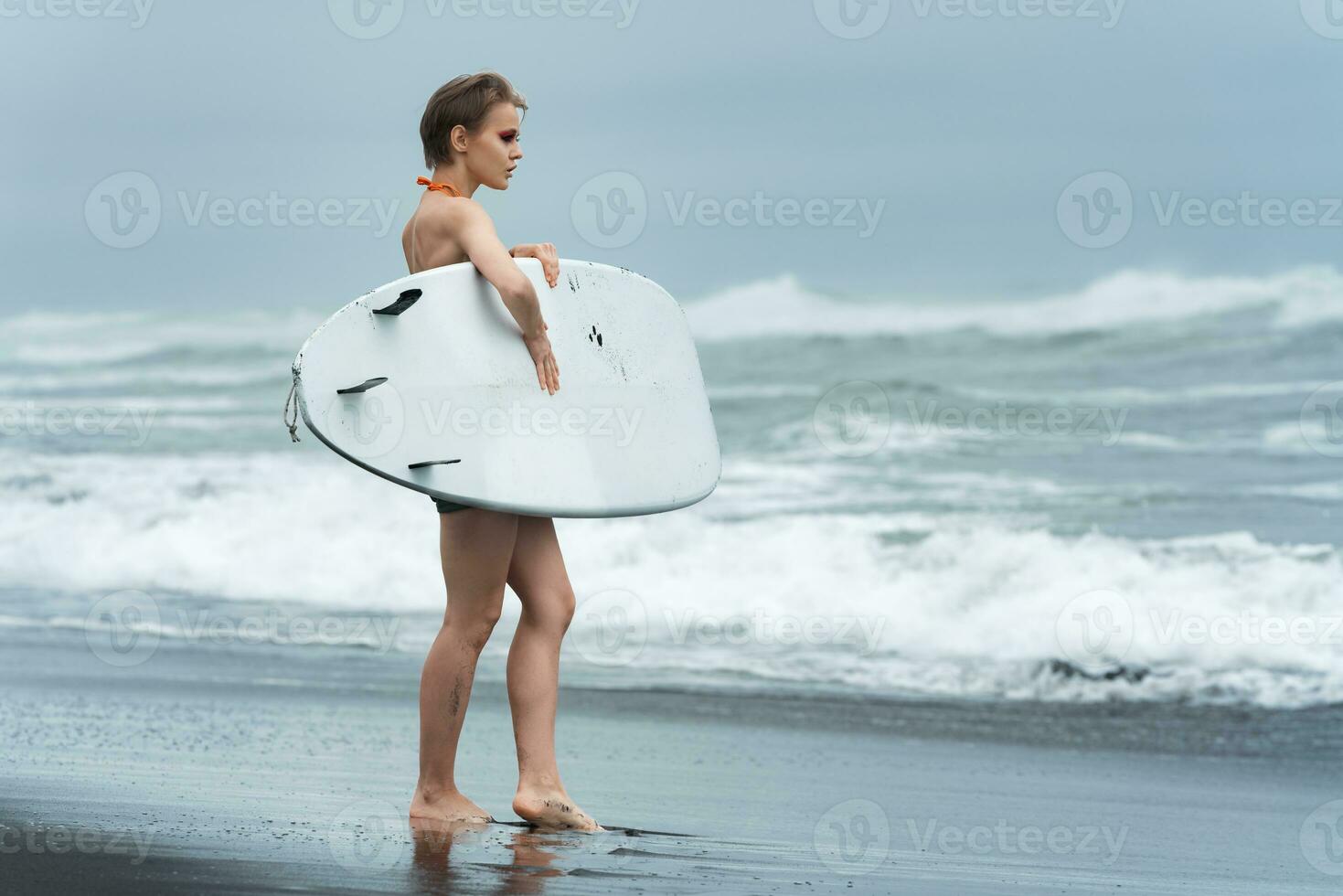 Surfer woman standing on sandy beach and carrying white surfboard against background of sea waves photo