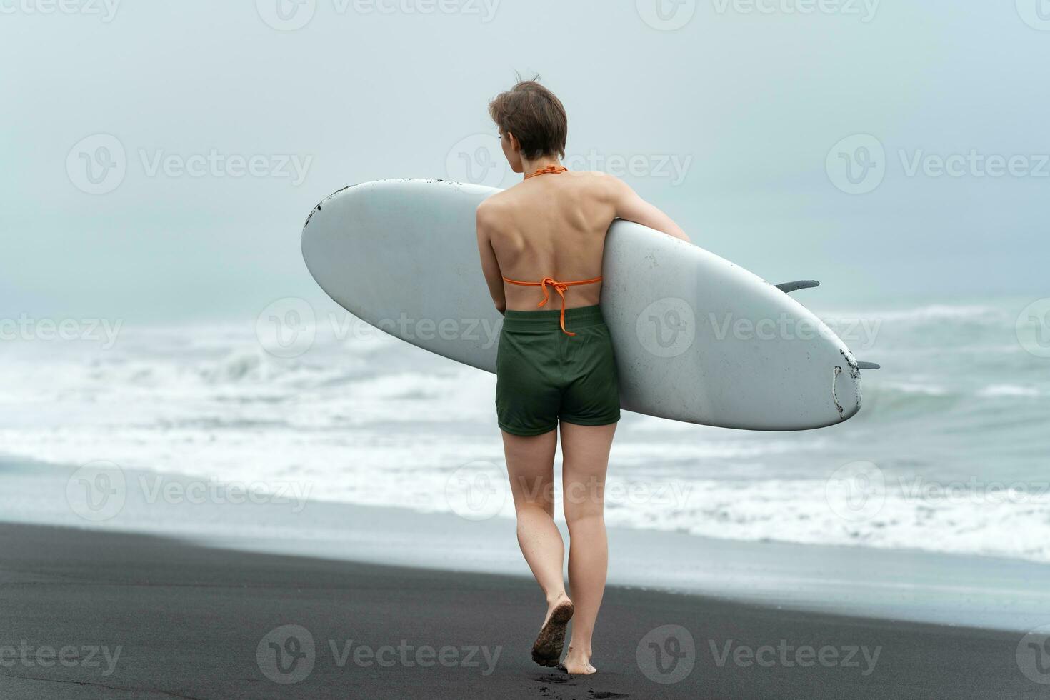 Rear view of unidentified female surfer walking on black sandy beach while carrying white surfboard photo