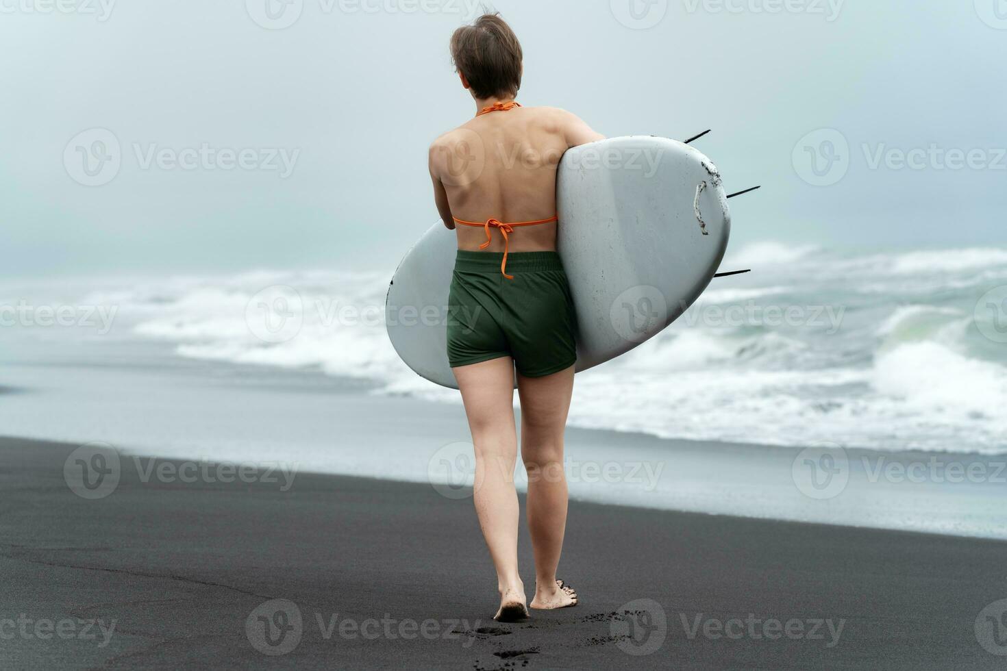 Rear view unrecognizable female surfer walking beach carrying surfboard on background of sea waves photo