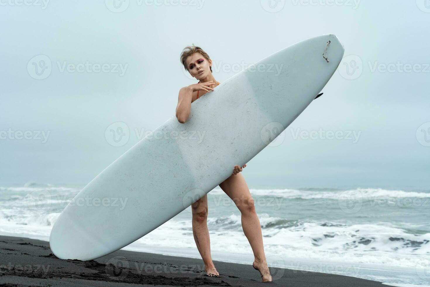 Woman surfer on beach holding surfboard and standing behind it and looking at camera photo