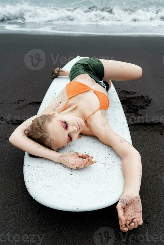 Female surfer is enjoying well deserved break on sandy beach, lying on surfboard with eyes closed photo