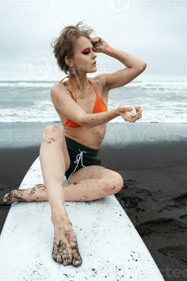 Sexy female surfer sitting on surfboard on sandy beach with sea waves in background photo