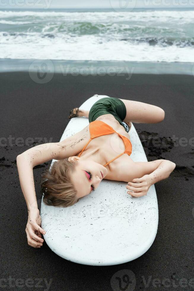 Female surfer is lying on surfboard on black sandy beach. With her eyes closed and in relaxed pose photo