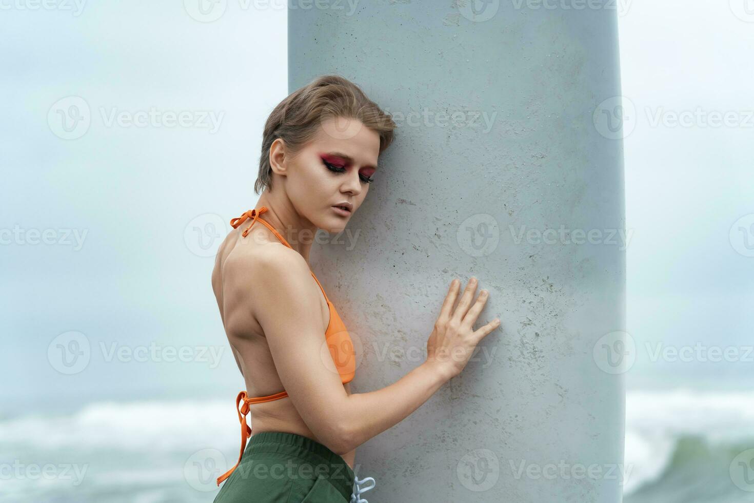 Female model with eyes closed holding surfboard and pressing it against herself in suggestive manner photo