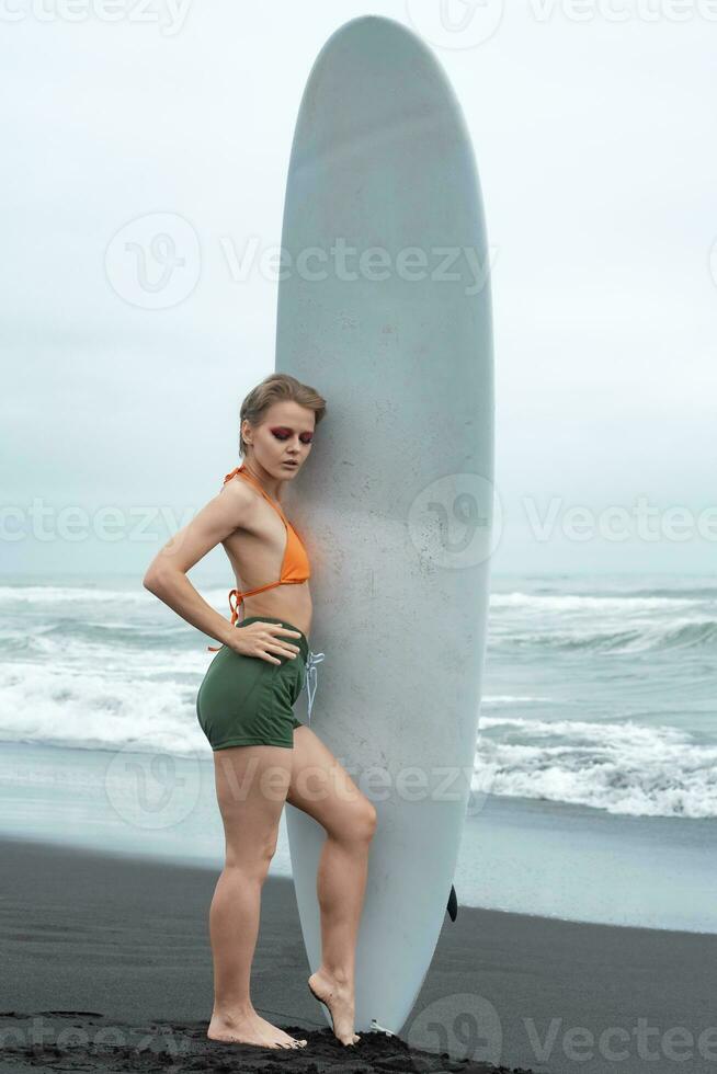 Female surfer stands on beach and holds white surfboard vertical on background of breaking waves photo