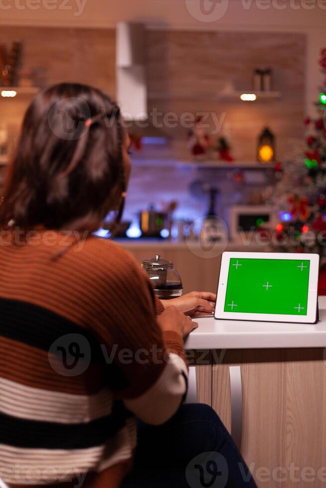 Caucasian adult watching green screen technology on tablet sitting at kitchen counter. Young woman using digital chroma key concept for background template, mockup gadget app on display photo
