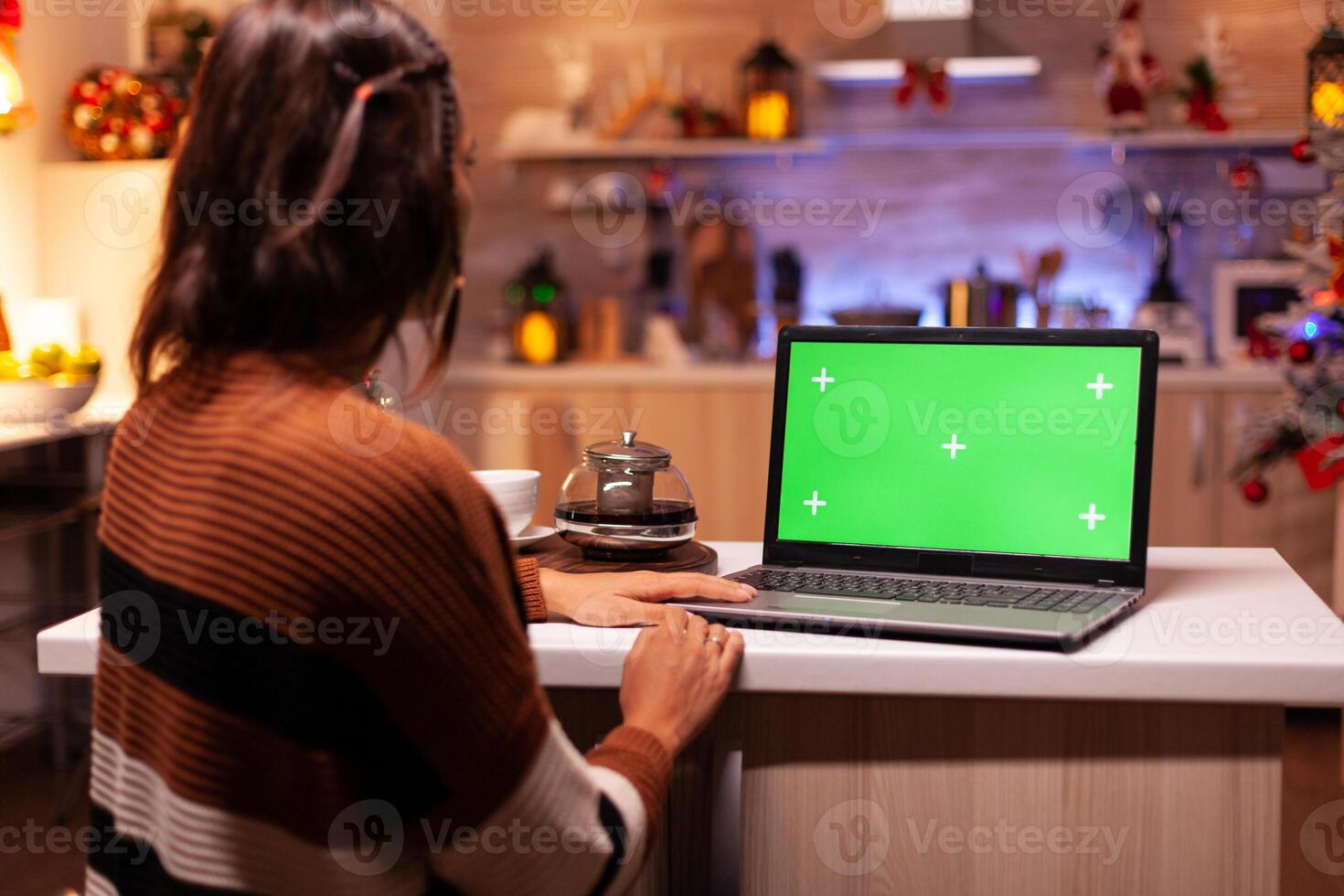 Modern young adult watching green screen on laptop digital device sitting at festive counter kitchen. Woman using computer chroma key gadget on background display for mockup template photo