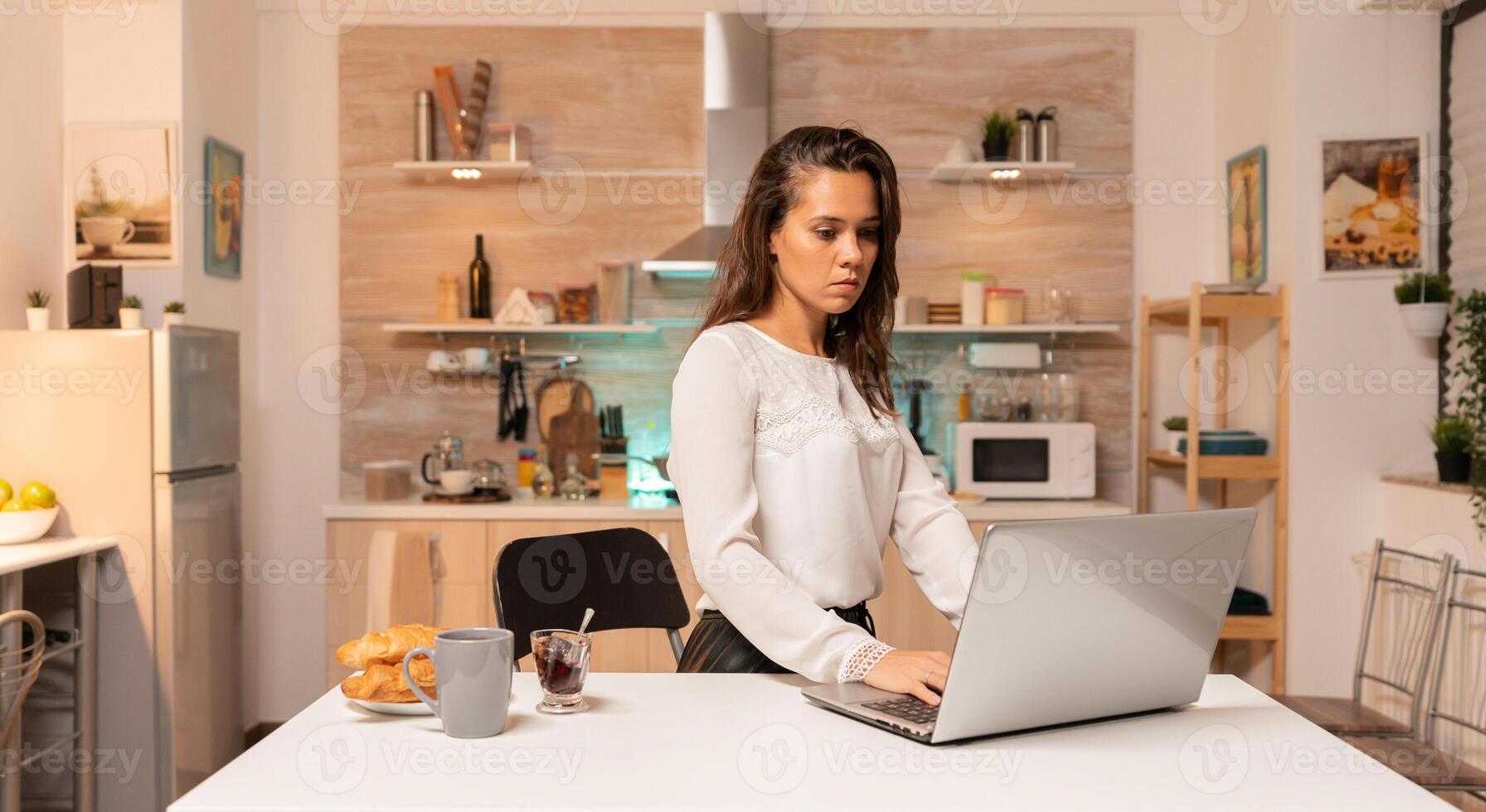 Tired businesswoman working overtime late at night using laptop in home office. Concentrated entrepreneur in home kitchen using notebook during late hours in the evening. photo