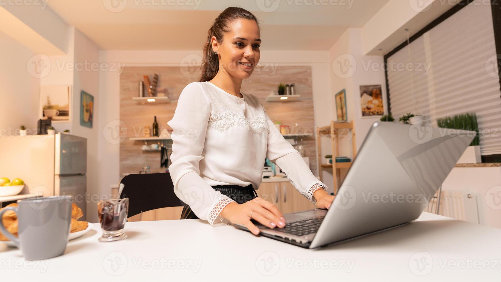 Busy businesswoman comunicating with potential client at night using laptop. Concentrated entrepreneur in home kitchen using notebook during late hours in the evening. photo
