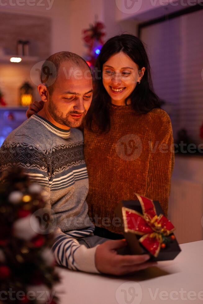 Husband surprising girlfriend with xmas present gift with ribbon on it celebrating christmas holiday in x-mas decorated kitchen. Happy joyful family enjoying winter season together photo