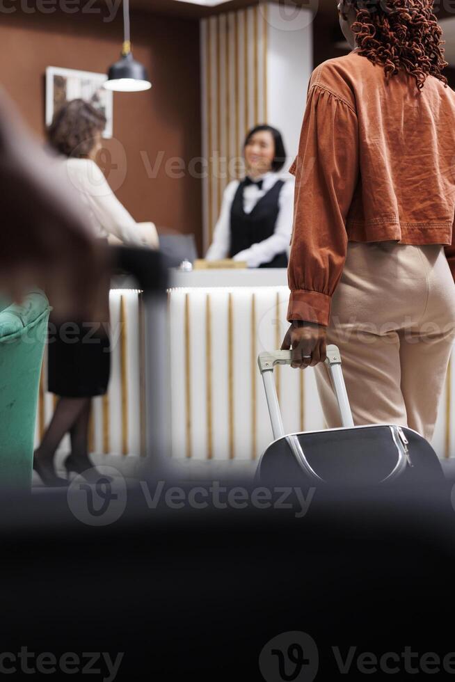Tourist waiting to register at hotel, carrying bags in lobby area with reception counter. Woman with luggage preparing to check in and ask about room accommodation at front desk. photo