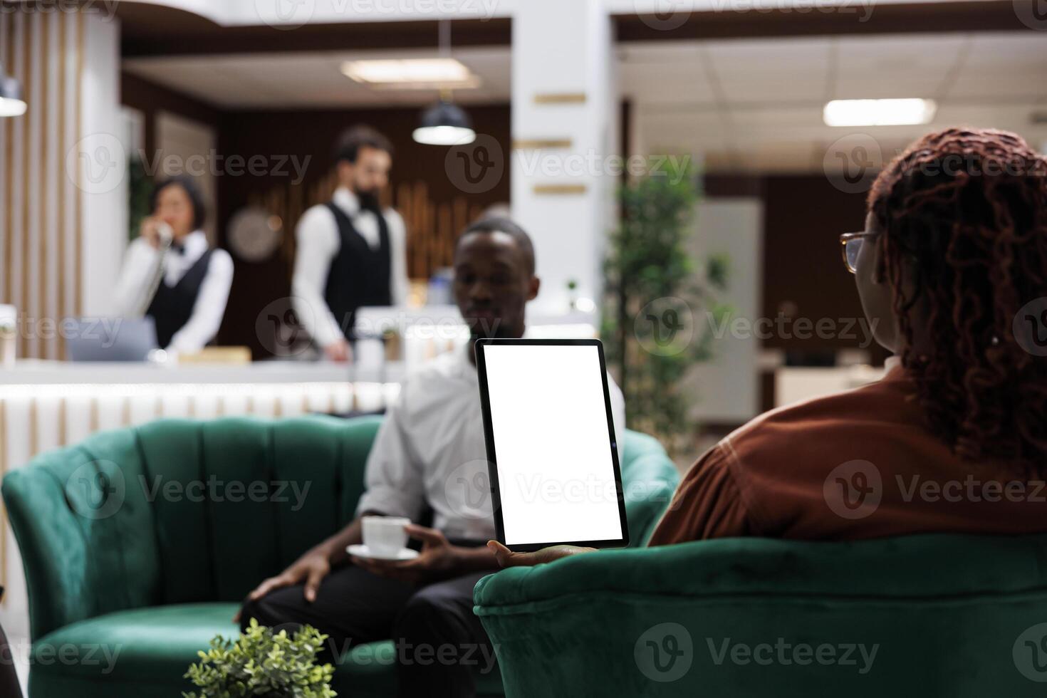 African american people at hotel using tablet with white screen, looking at blank template display and sitting in lounge area. Female guest holding device with isolated copyspace. photo