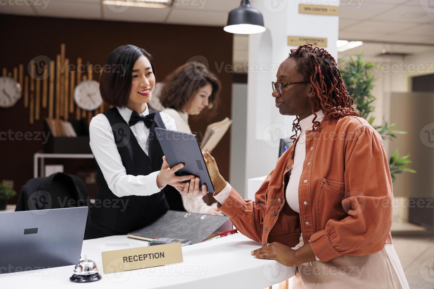 Young woman checking in at luxury hotel, filling in registration forms for records in lobby. African american guest doing check in with female receptionist at front desk, booking files. photo