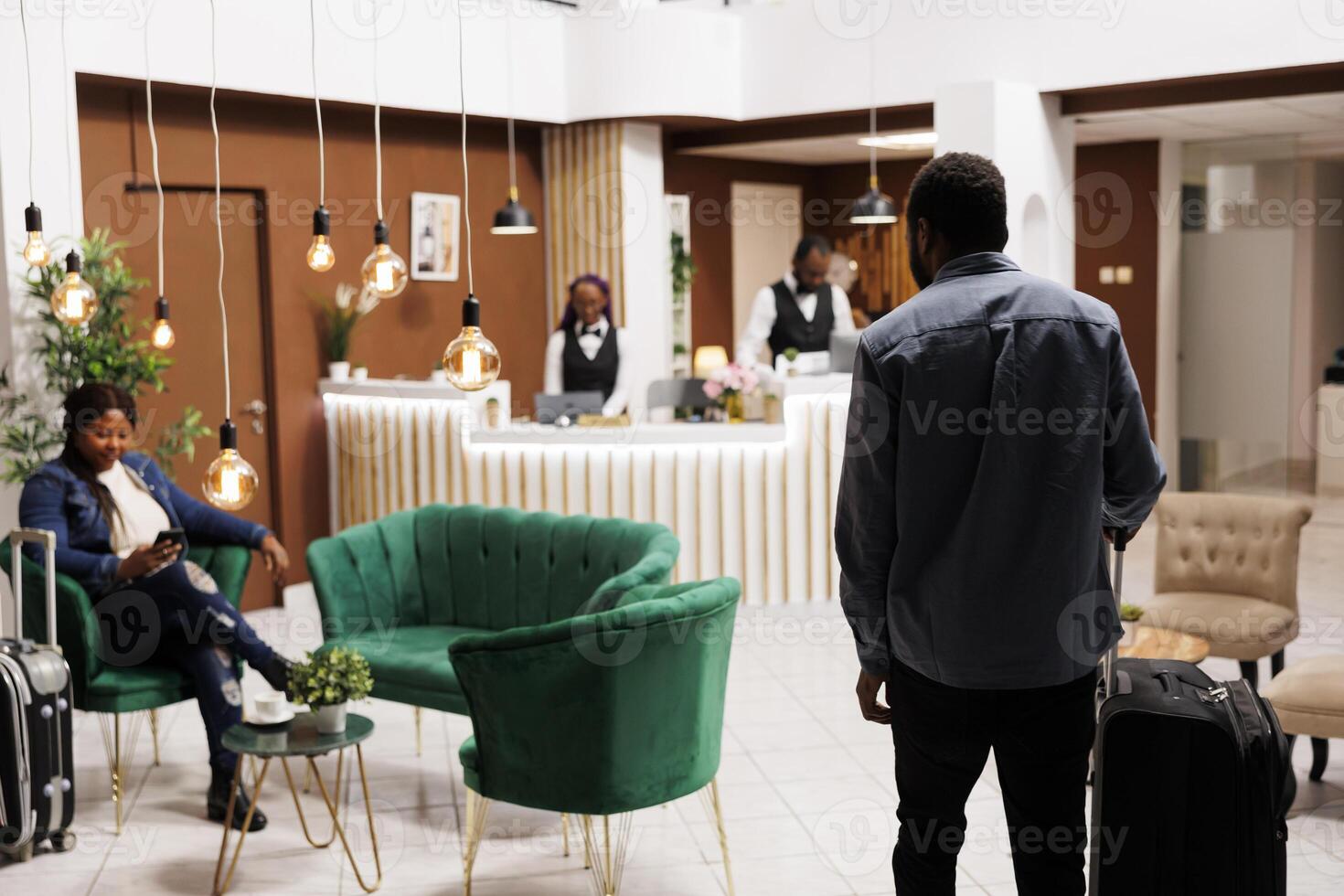 African American man tourist arriving at hotel, standing with luggage in foyer or lobby, rear view. People tourist arrive at resort waiting for check-in procedure, holiday season photo