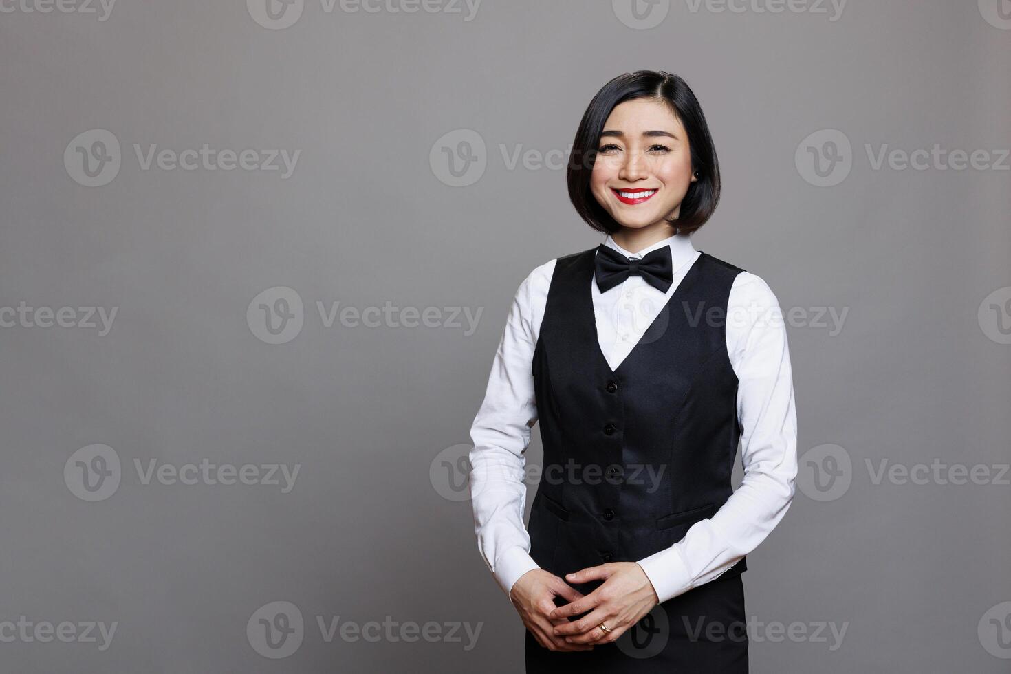 Attractive smiling asian receptionist wearing professional uniform standing and looking at camera with positive facial expression. Young waitress studio portrait on gray background photo