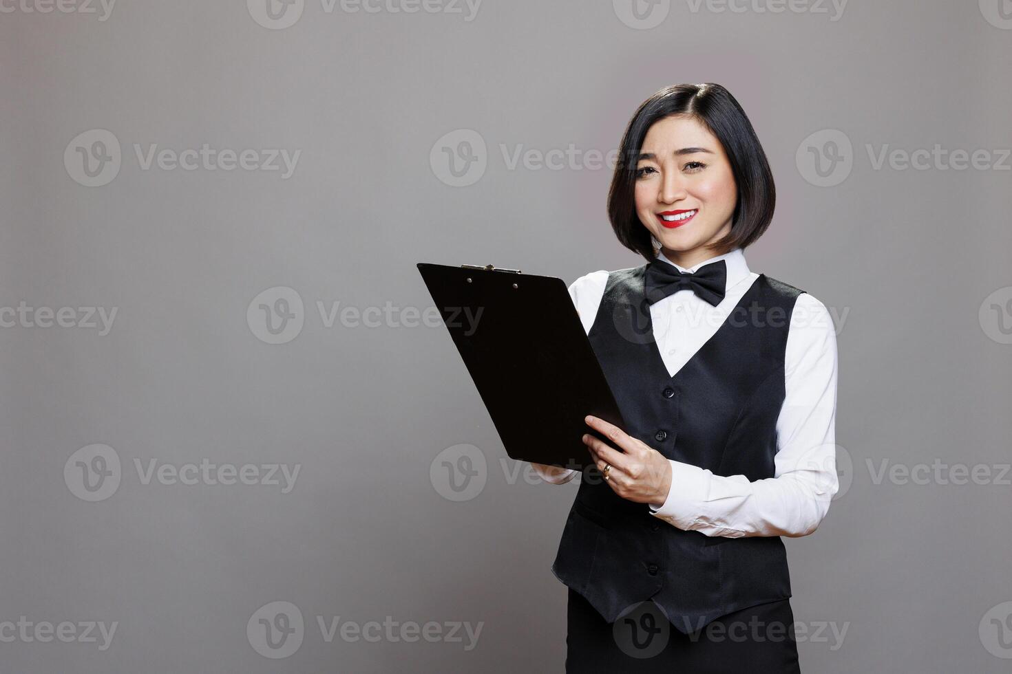 Attractive smiling asian waitress writing on a clipboard and looking at camera with positive facial expression. Cheerful young receptionist in uniform taking notes while posing in studio portrait photo