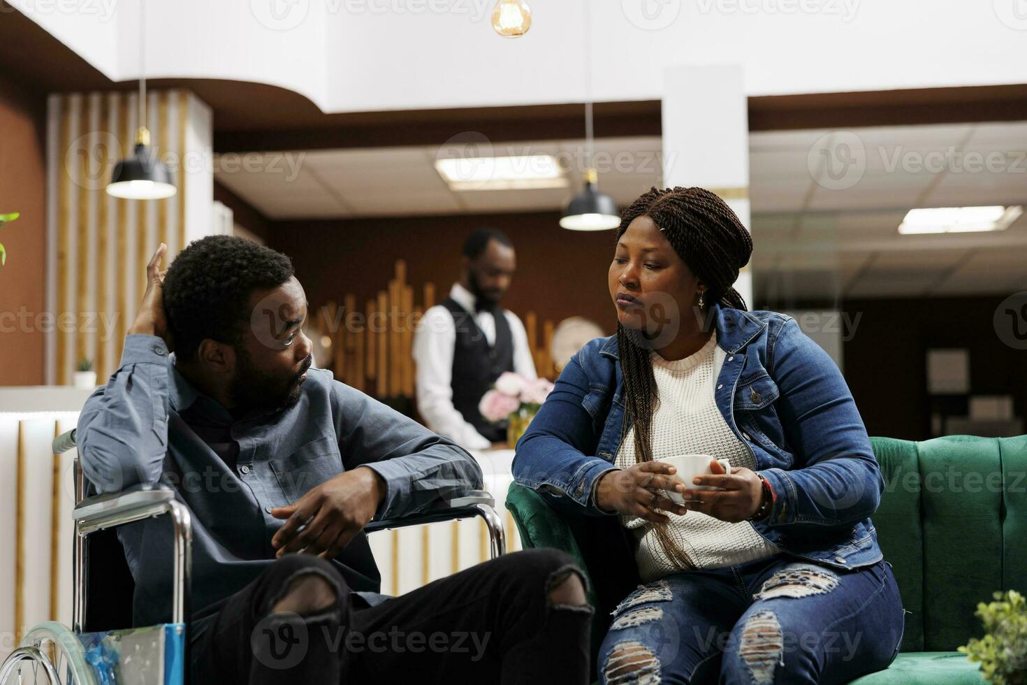 Handicapped travel. Young African American man on wheelchair waiting with wife in hotel lobby, guest with mobility impairments, traveling with disability. Travel companions for disabled travelers photo