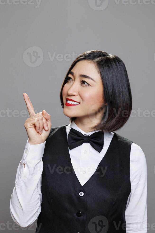 Smiling asian woman receptionist showing upwards and looking to side while advertising product. Cafe cheerful waitress wearing professional uniform pointing up with finger photo