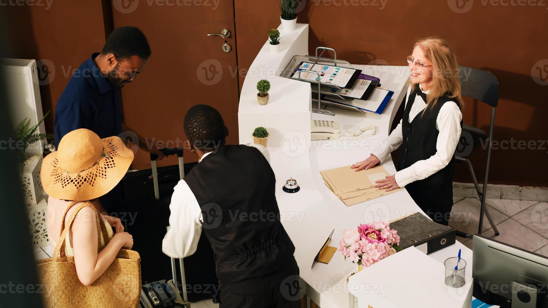 People arriving at hotel front desk, preparing to do check in at reception counter and see room reservation. Guests on honeymoon during summertime talking to concierge in lobby. Handheld shot. photo
