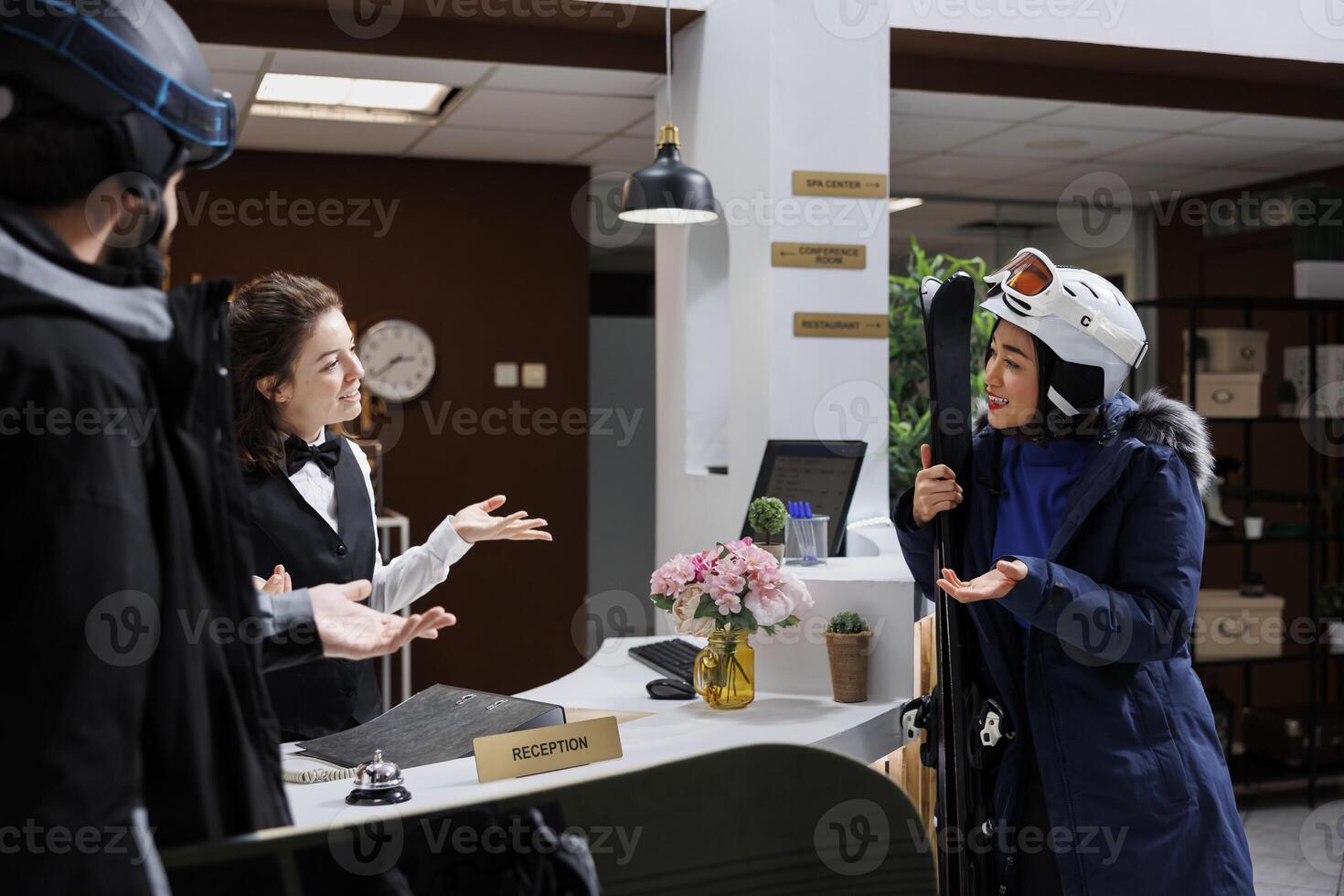 Friendly receptionist assisting female tourist with reservation and information on best skiing slopes at winter mountain resort. Young travelers carrying wintersports equipment talking with concierge. photo