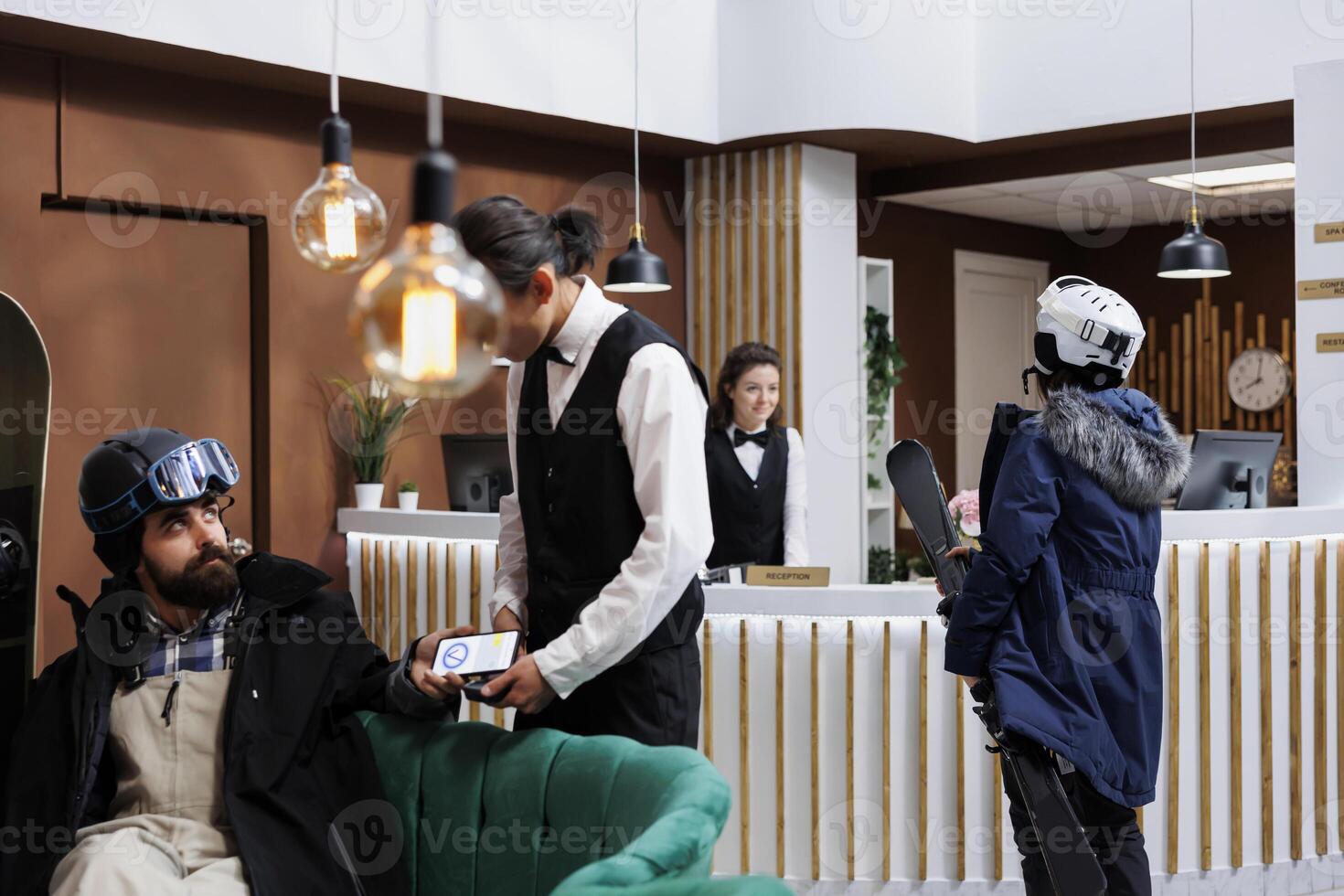 Caucasian man in winter clothing using mobile device on pos machine provided by male employee at hotel reception. Woman carrying skiing skis approaches front desk for check-in at ski mountain resort. photo