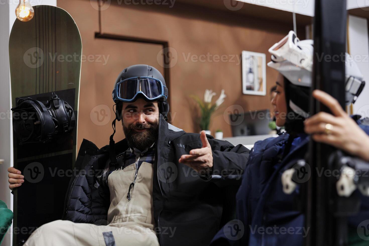 Youthful pair outfitted in cold-weather attire seated on sofa situated in hotel lobby engaged in conversation. Happy man and lady are clutching their wintersports gear set for snowy escapade. photo