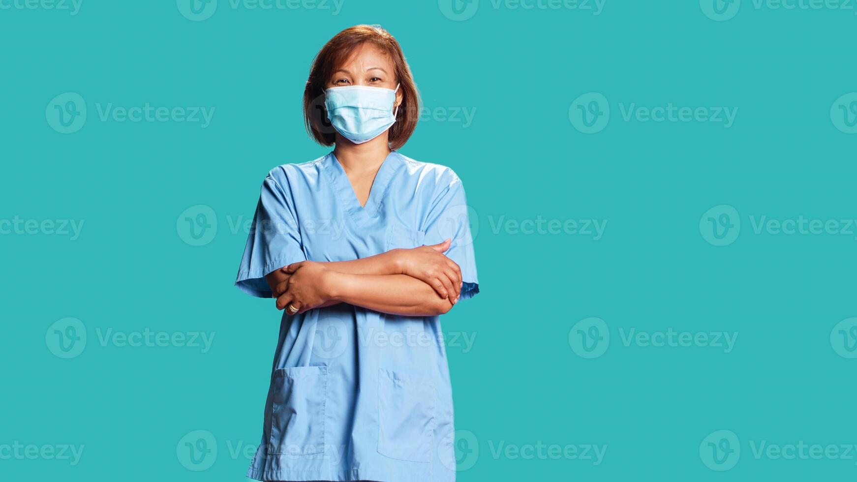 Upbeat smiling woman wearing protective clinical face mask to stop bacteria spreading. Portrait of lively nurse in professional scrubs with arms folded, isolated over blue studio background photo