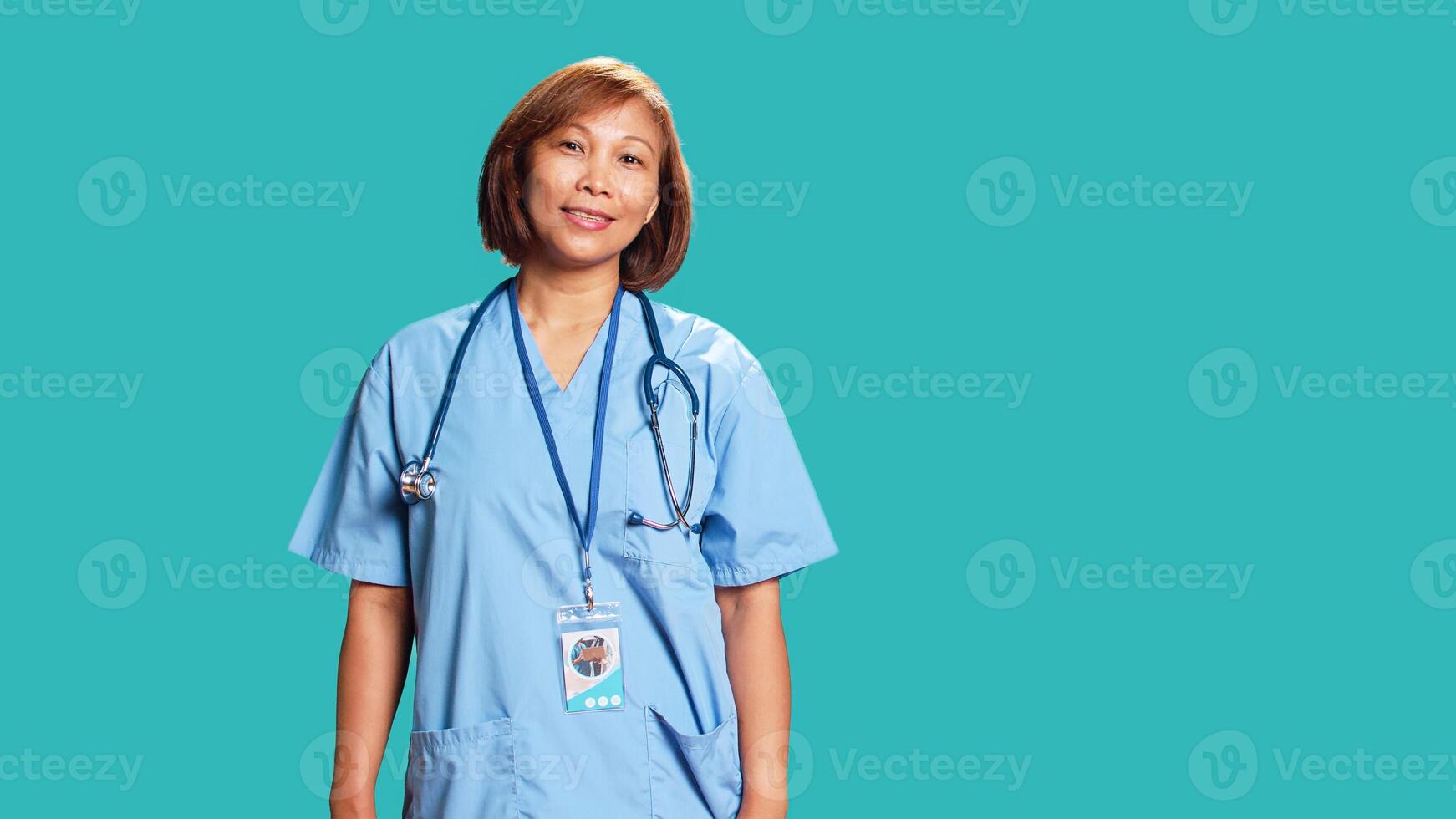 Happy proficient BIPOC nurse smiling while looking at camera. Portrait of cheerful asian healthcare specialist wearing professional scrubs, isolated over blue studio background photo