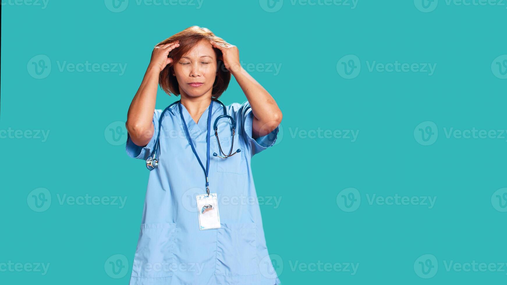 Overworked nurse suffering from migraine pain after exhausting day at work. Healthcare specialist isolated over blue studio background affected by troubling headache during tiresome job shift photo