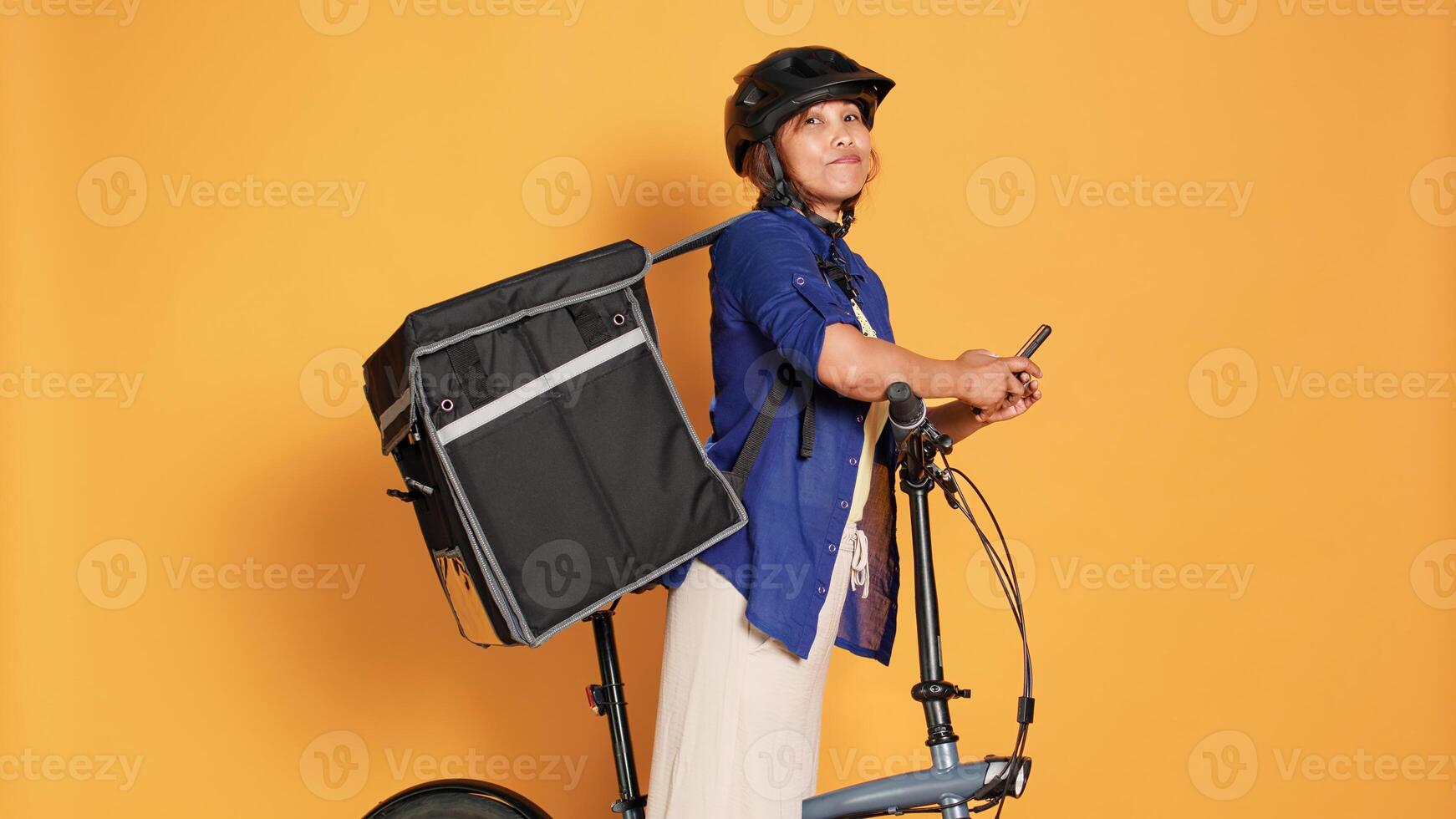 Impatient courier looking at phone, waiting to receive next delivery location. Asian cyclist preparing to pick up takeaway food from restaurant to customer, isolated over orange studio background photo