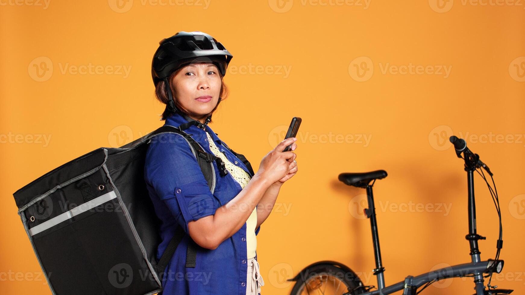 Courier delivering food, texting customer to pick up order. Asian woman in a rush, upset with client taking long time to get purchased meal, isolated over orange studio background photo