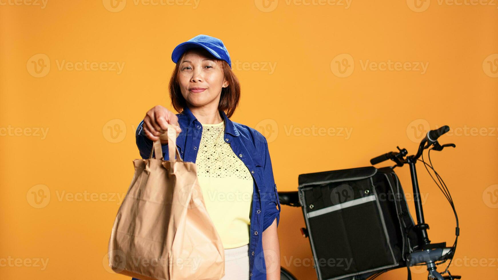 Slow motion portrait shot of friendly courier handing lunch bag to customer, showing thumbs up sign. Food delivery cyclist providing meal order to client, isolated over studio background photo