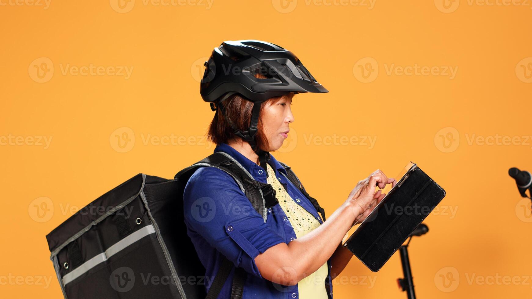 Part time delivery worker typing customer order details on tablet, close up. Asian courier wearing safety helmet and food backpack checking online app, isolated over orange studio background photo