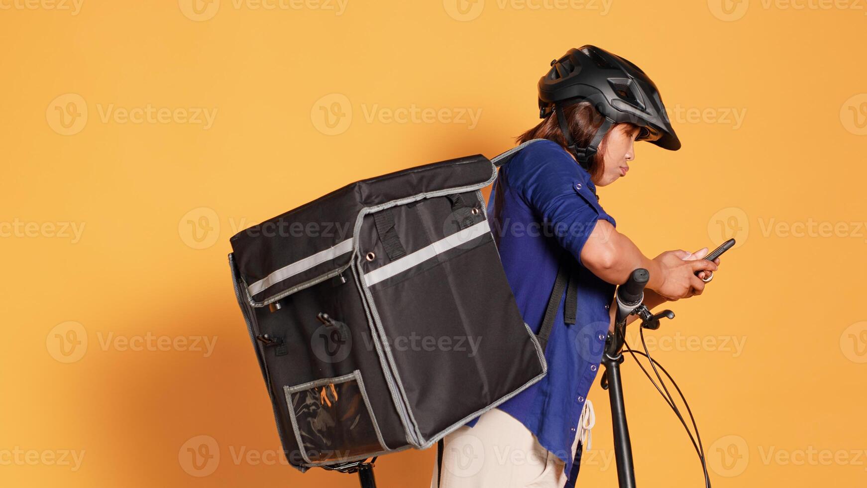 Handheld camera shot of courier texting client after delivering wrong takeout order. Bike rider wearing safety helmet and thermic bag holding food, isolated over studio background photo