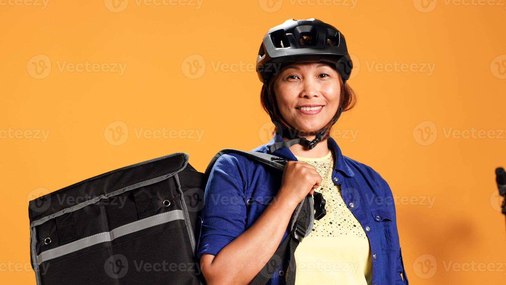 Portrait of happy asian courier using bike and thermic bag to transport perishables to customer. Delivery part time employee isolated over orange studio background wearing safety gear, close up photo