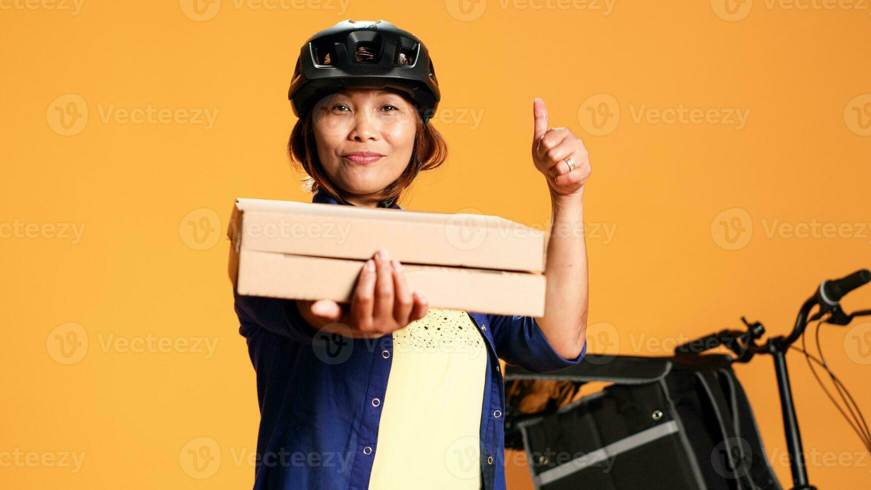 Close up portrait of happy professional courier pointing to takeout pizza boxes. Friendly bike rider isolated over orange studio background showing approval gesture while delivering fast food meals photo