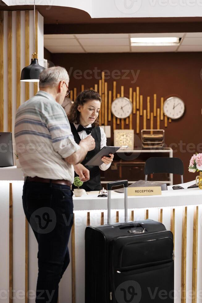 Elderly male traveller checks in at registration counter of hotel lobby, eager to begin enjoyable holiday. Retired senior man fills out reservation documents with help from kind female receptionist. photo