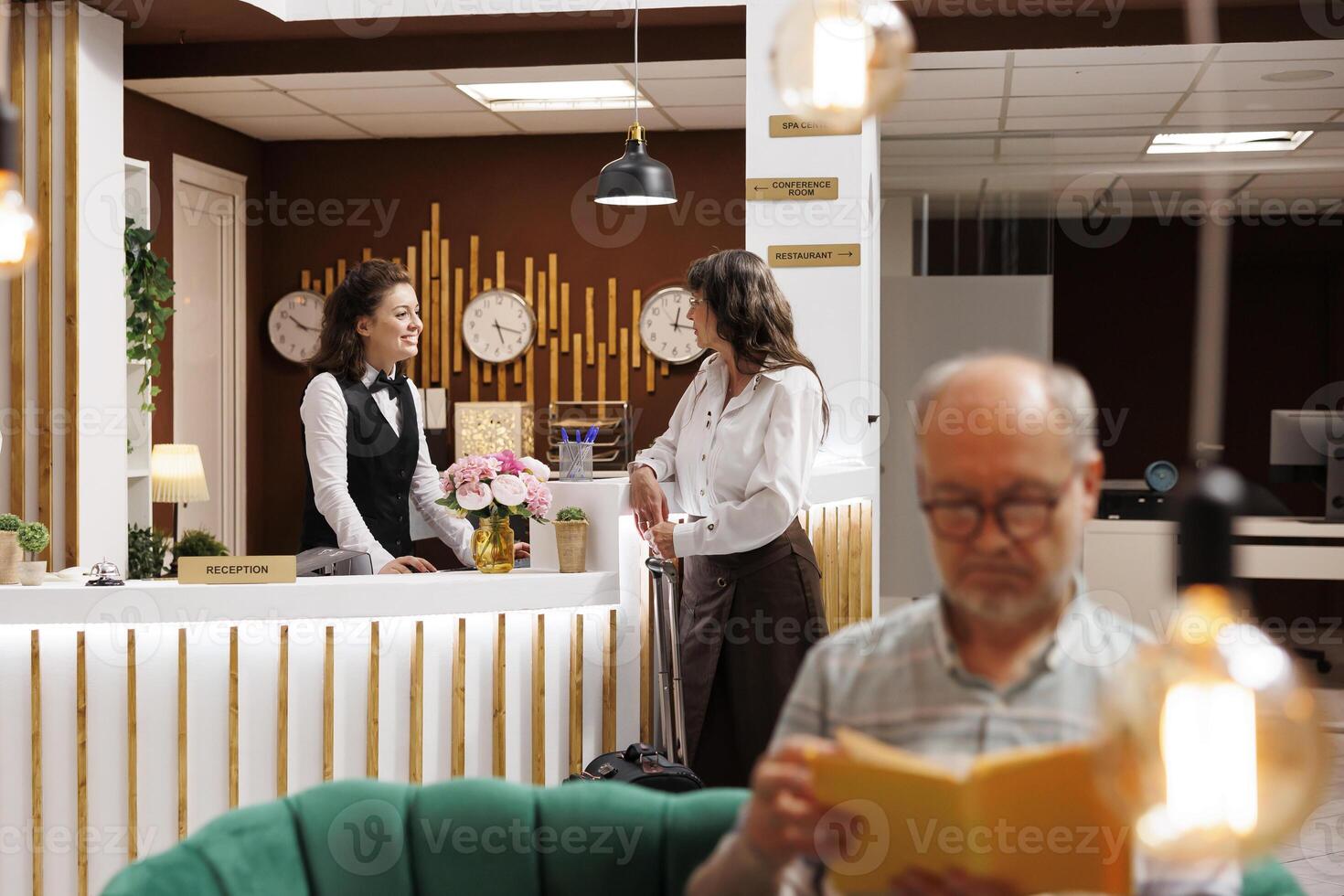 Elderly female guest with luggage speaking to young receptionist at front desk of luxury resort. Old man with a novel relaxes in hotel lobby while senior woman does booking procedure at counter. photo