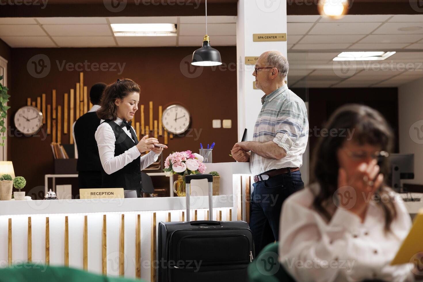 Helpful female concierge assists an elderly male guest with registration at hotel reception counter. Senior man waits at front desk while receptionist verifies identity with his passport for check in. photo