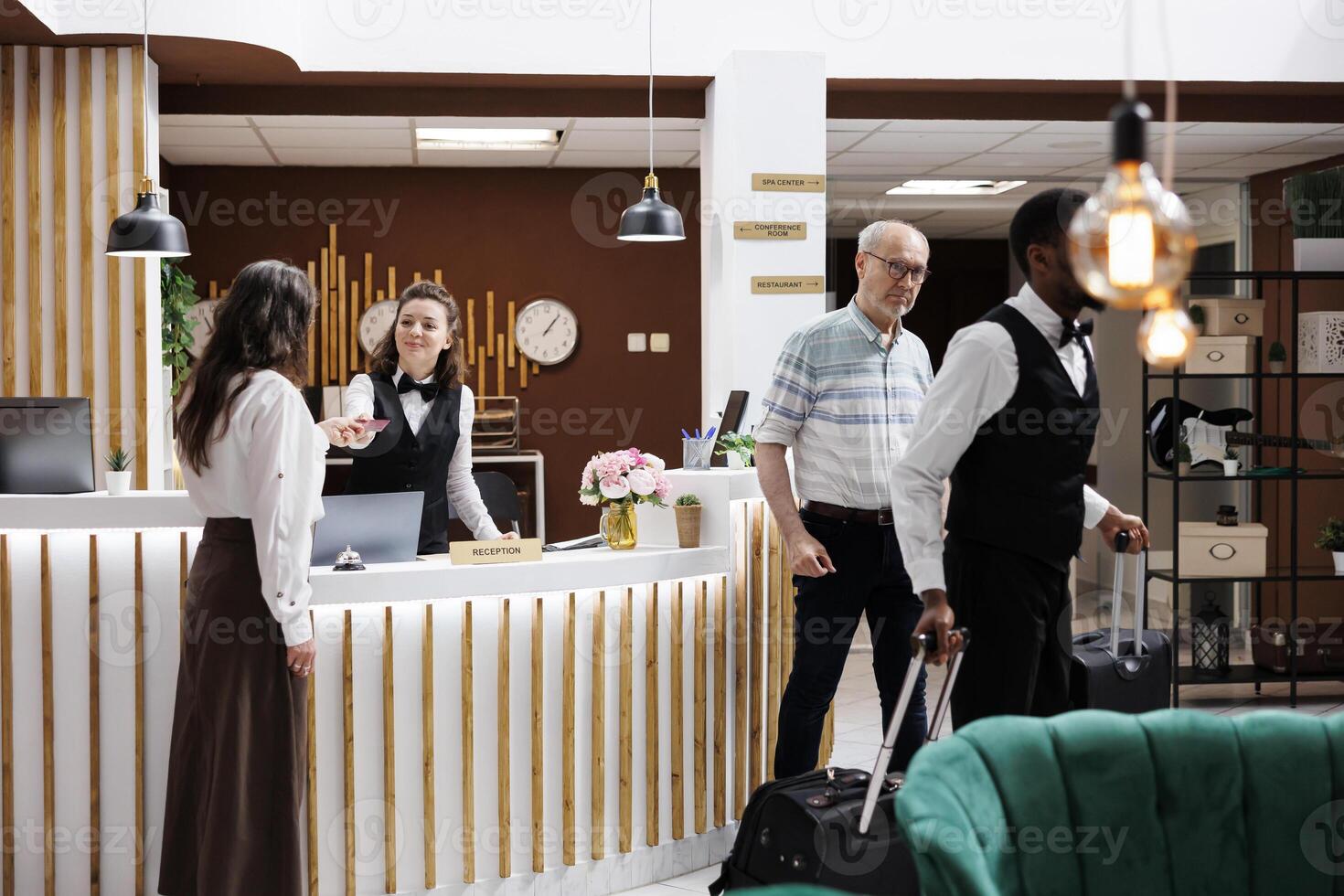 Female concierge assists elderly lady at front desk, providing smooth check-in process and verifying reservations. African american bellboy helps retired senior man with suitcases to hotel room. photo