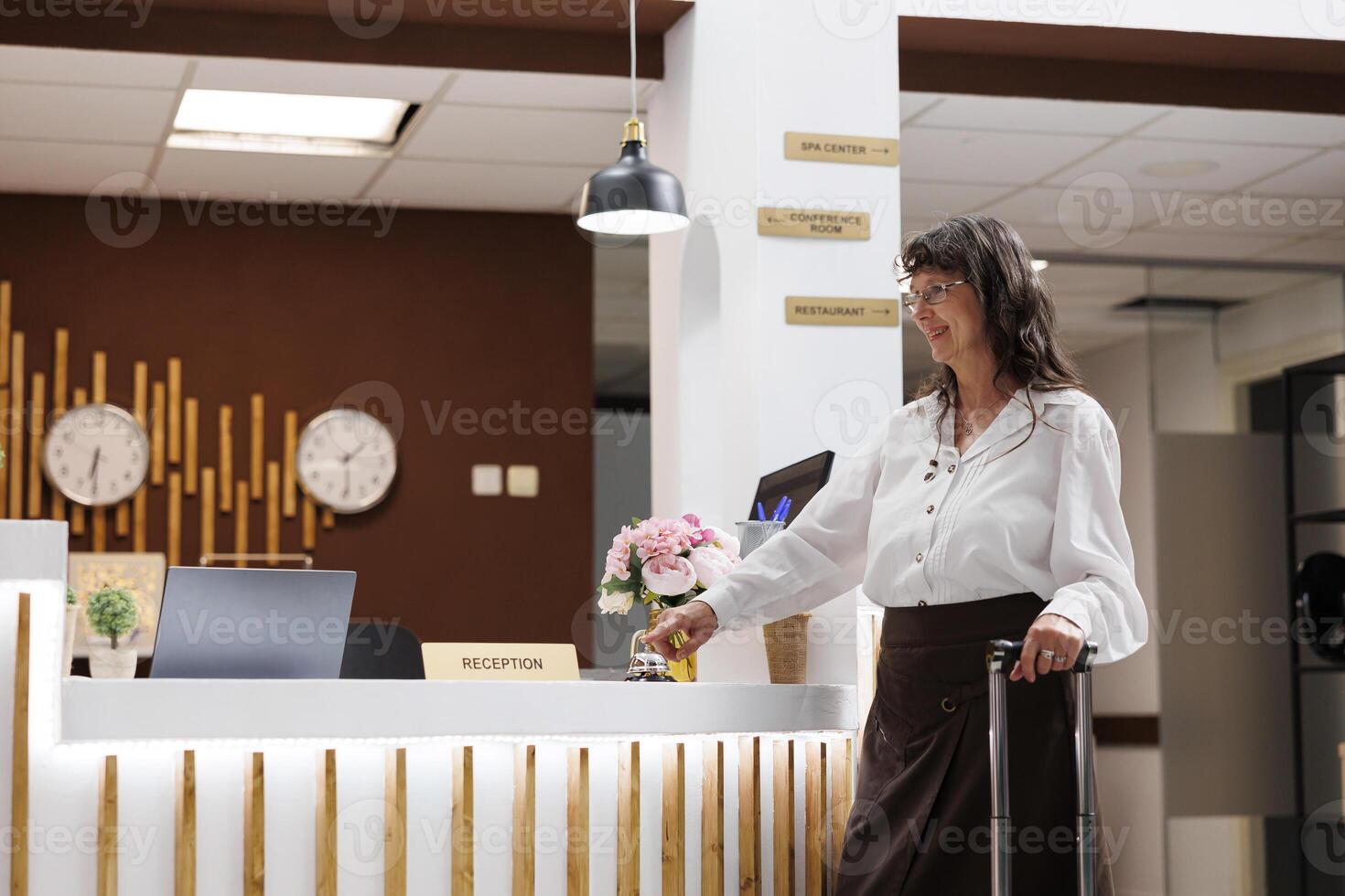 Senior woman reaches registration counter and rings the metallic service bell asking for services from hotel receptionists. Caucasian elderly female tourist awaiting assistance from resort personnel. photo