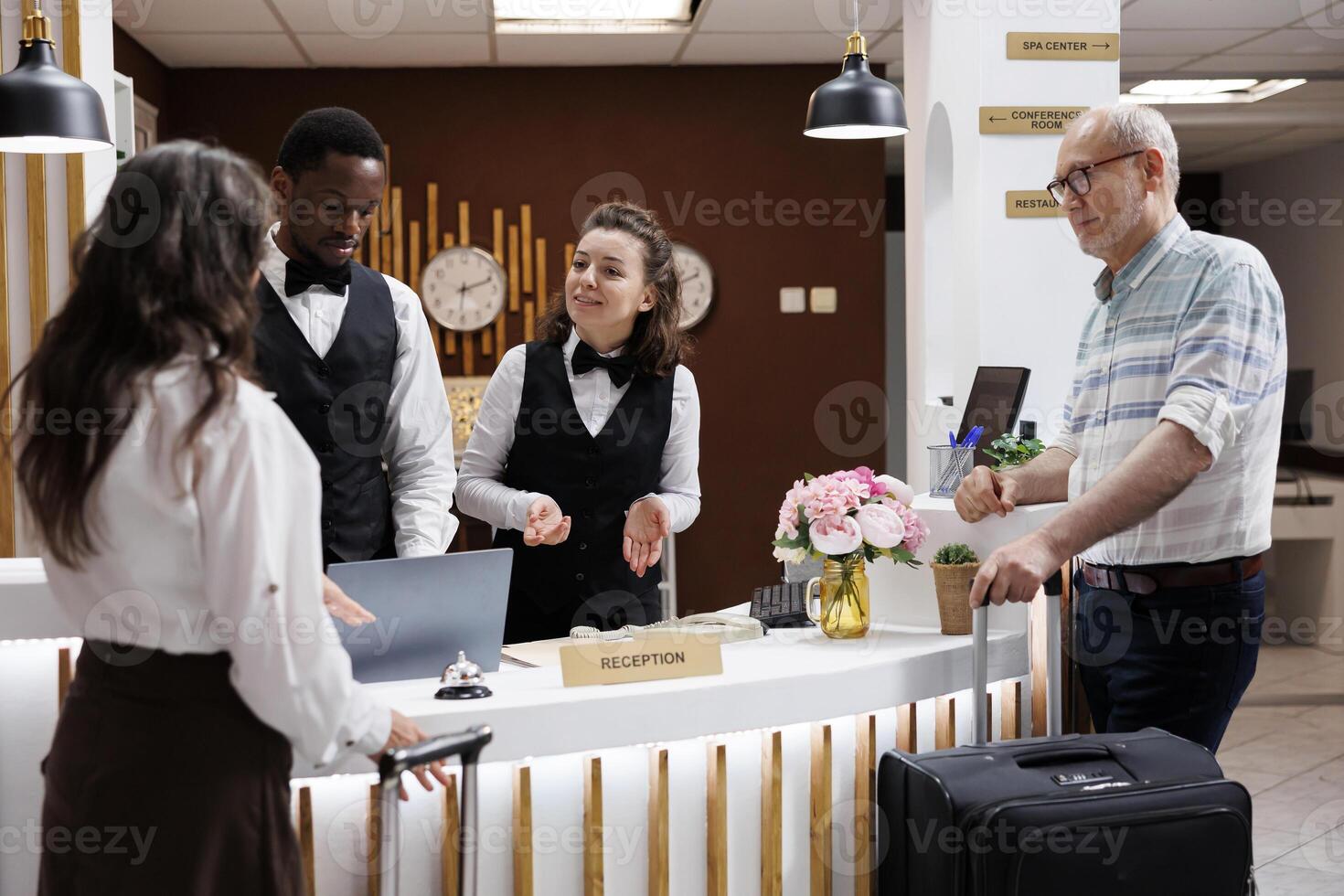 Assisting elderly guests at upscale hotel reception, concierge welcomes visitors with warmth and efficiency. Front desk staff ensures smooth check-in process for senior visitors in elegant lobby. photo