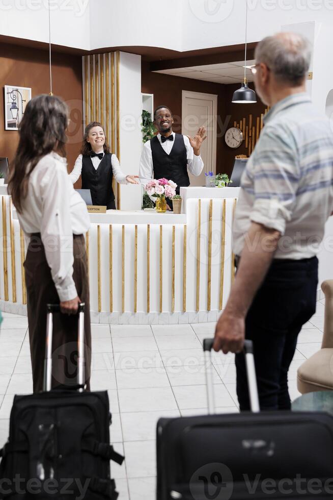 Caucasian elderly couple enters hotel and approaches front desk for reservation assistance from multiethnic employees. Professional receptionists greeting retired seniors with smiles. photo