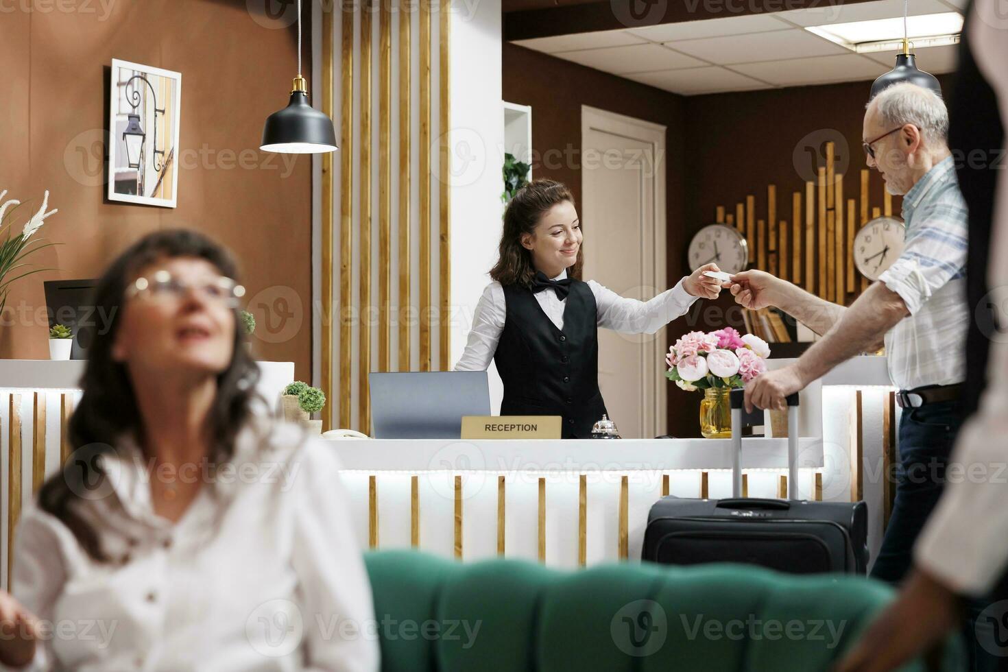 Arriving at front desk, old man checks-in hotel reception, assisted with registration and given room key. Retired senior male tourist receives access card while bellboy helps with guest in lounge area. photo