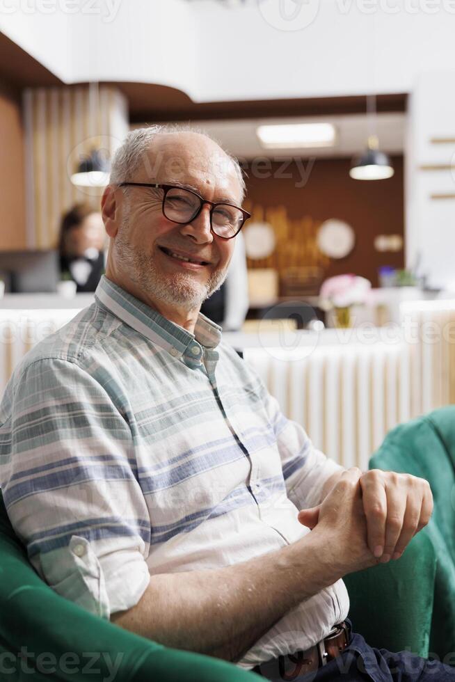 Detailed view of smiling elderly male tourist sitting in exclusive lounge area of resort. Close-up of caucasian senior man looking at camera while waiting for check-in in hotel reception. photo