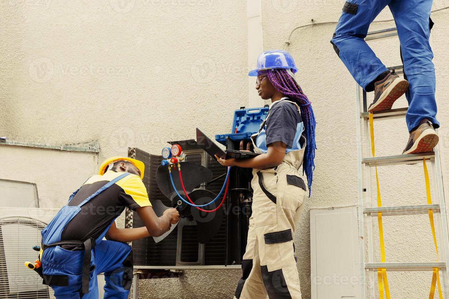 Seasoned mechanic cleaning and lubricating hvac system internal parts, checking refrigerant level and calibrating thermostats while african american colleague checks maintenance plan on laptop photo