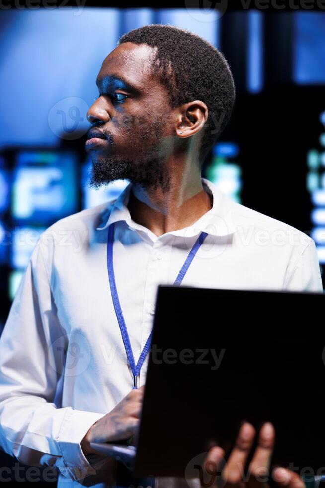 African american manager in server hub using laptop to find firewall misconfigurations affecting rackmounts performance and connectivity, leading to sluggish data transfer rates photo